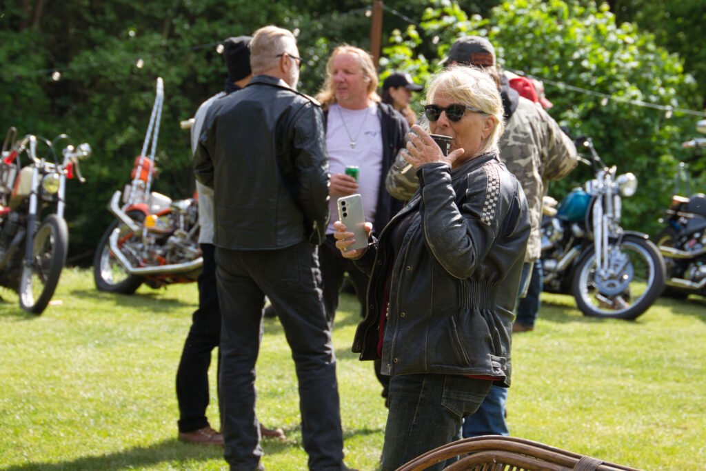 Eine Frau mit Sonnenbrille und Lederjacke lächelt und hält ein Smartphone in der Hand, umgeben von Menschen und geparkten Motorrädern auf einer Wiese mit Bäumen im Hintergrund.