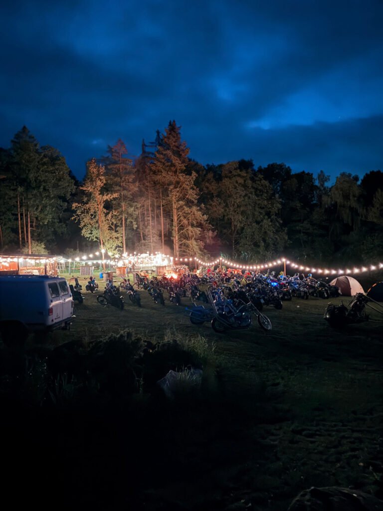 Eine nächtliche Szene im Freien mit Reihen von geparkten Motorrädern, Lichterketten und Bäumen im Hintergrund. Menschen versammeln sich in der Nähe von hell erleuchteten Essensständen unter einem dunkelblauen Himmel.