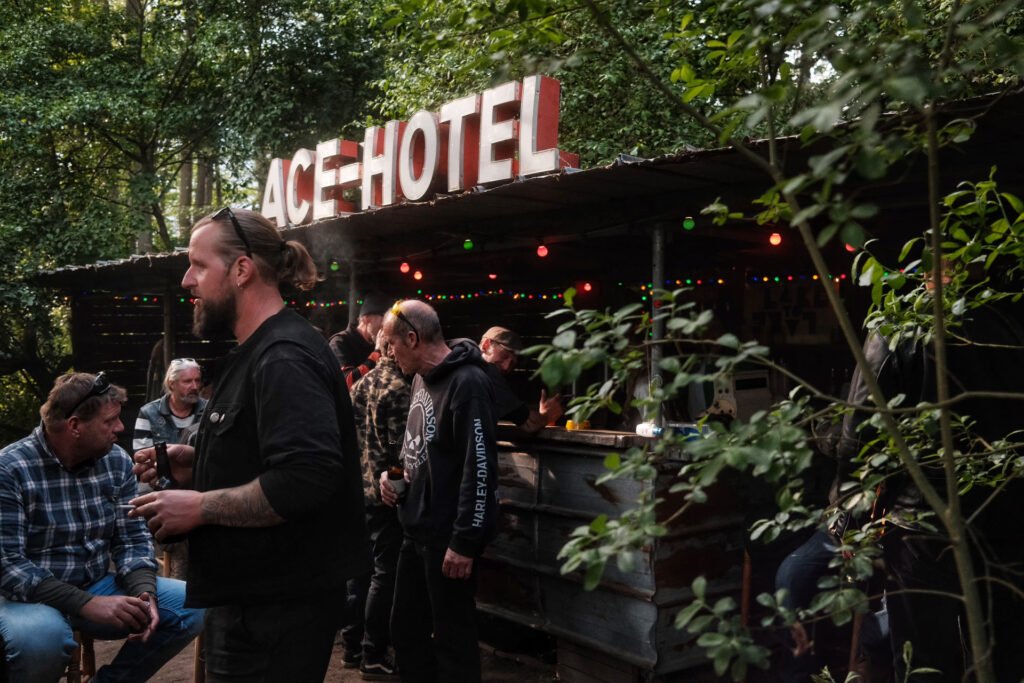 Eine Gruppe von Menschen trifft sich vor einer rustikalen Bar mit einem großen Schild "ACE HOTEL". Bunte Lichterketten hängen darüber, und Bäume umgeben die lebhafte Umgebung.