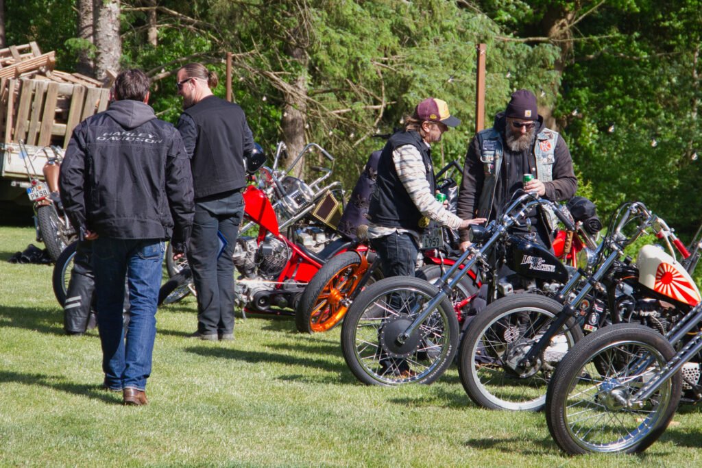 Vier Personen stehen draußen im Gras in der Nähe von Custom-Motorrädern, unterhalten sich und sehen sich die Motorräder an. Im Hintergrund sind Bäume und ein Stapel Holzpaletten zu sehen. Die Gruppe ist mit lässigen Jacken und Hüten bekleidet.