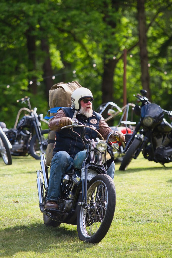 Ein älterer Mann mit weißem Bart, der einen Helm und eine Sonnenbrille trägt, fährt auf einem Oldtimer-Motorrad mit einem großen Rucksack über eine Wiese, mit anderen Motorrädern und Bäumen im Hintergrund.