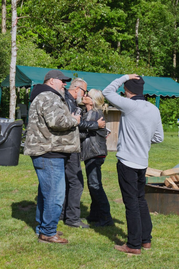 Vier Erwachsene stehen draußen auf grünem Gras, unterhalten sich und tauschen sich aus, in der Nähe eines Zeltdaches und von Feuerholz. Zwei Personen umarmen sich, während eine andere mit einer Hand auf dem Kopf steht. Im Hintergrund sind Bäume und Grünflächen zu sehen.