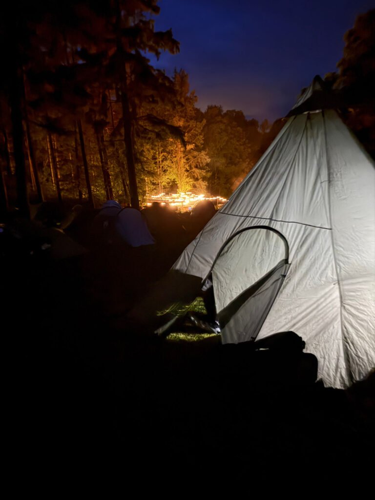Ein weißes Zelt wird bei Nacht in einem dunklen Wald beleuchtet. Warme Lichter leuchten im Hintergrund zwischen den Bäumen und schaffen eine gemütliche Zeltplatzatmosphäre unter einem tiefblauen Himmel.