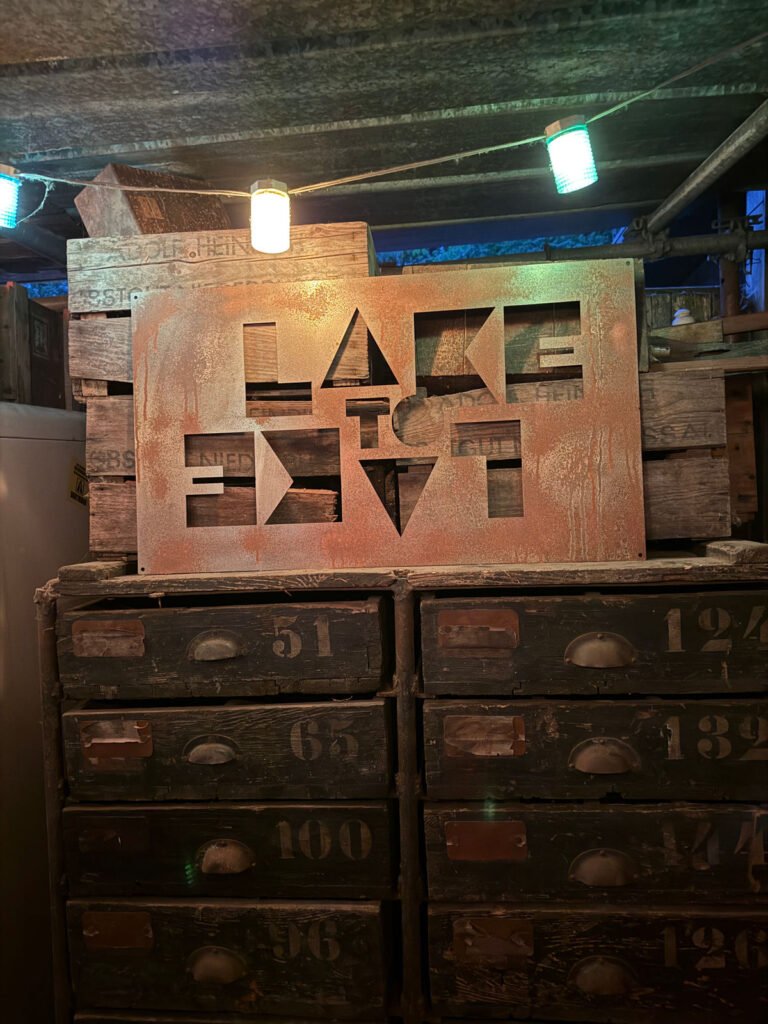 Ein Metallschild mit ausgeschnittenen Buchstaben, auf dem "LACE TO EXIT" steht, befindet sich auf alten, verwitterten Holzschubladen und -kisten in einer schwach beleuchteten, rustikalen Umgebung, über der grüne und blaue Lichter hängen.