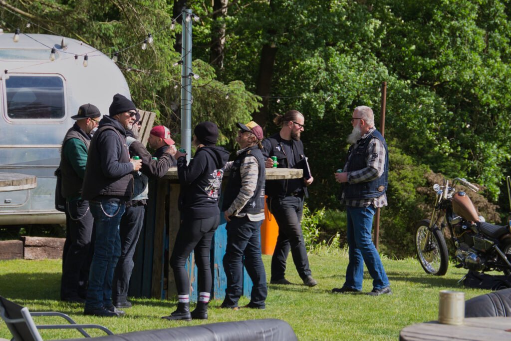 Eine Gruppe von Menschen mit Jacken, Hüten und Sonnenbrillen steht an einem sonnigen Tag um einen hohen Tisch im Freien in der Nähe eines Oldtimer-Anhängers herum und unterhält sich, während im Hintergrund ein Motorrad und Bäume zu sehen sind.