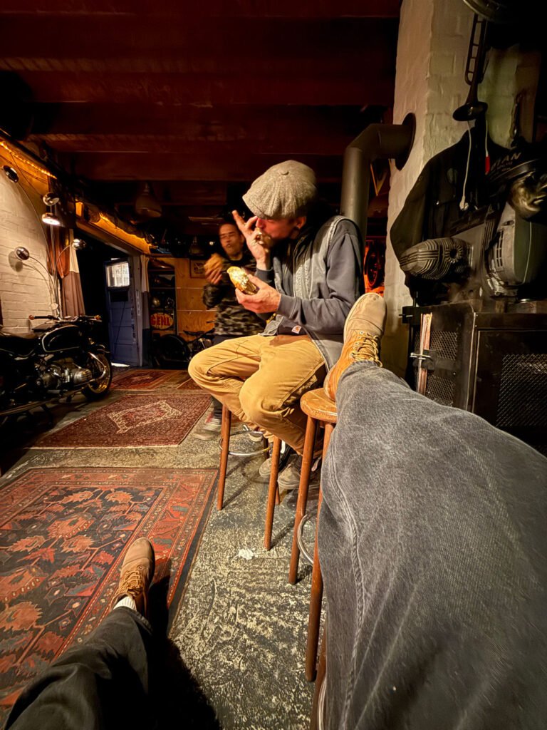 Ein Mann mit einer Mütze sitzt auf einem Hocker und isst, wobei er mit erhobenem Finger gestikuliert. Eine andere Person steht hinter ihm. Die Szene befindet sich in einem Innenraum mit warmer Beleuchtung, einem Motorrad und Teppichen. Die Beine des Fotografen sind im Vordergrund zu sehen.