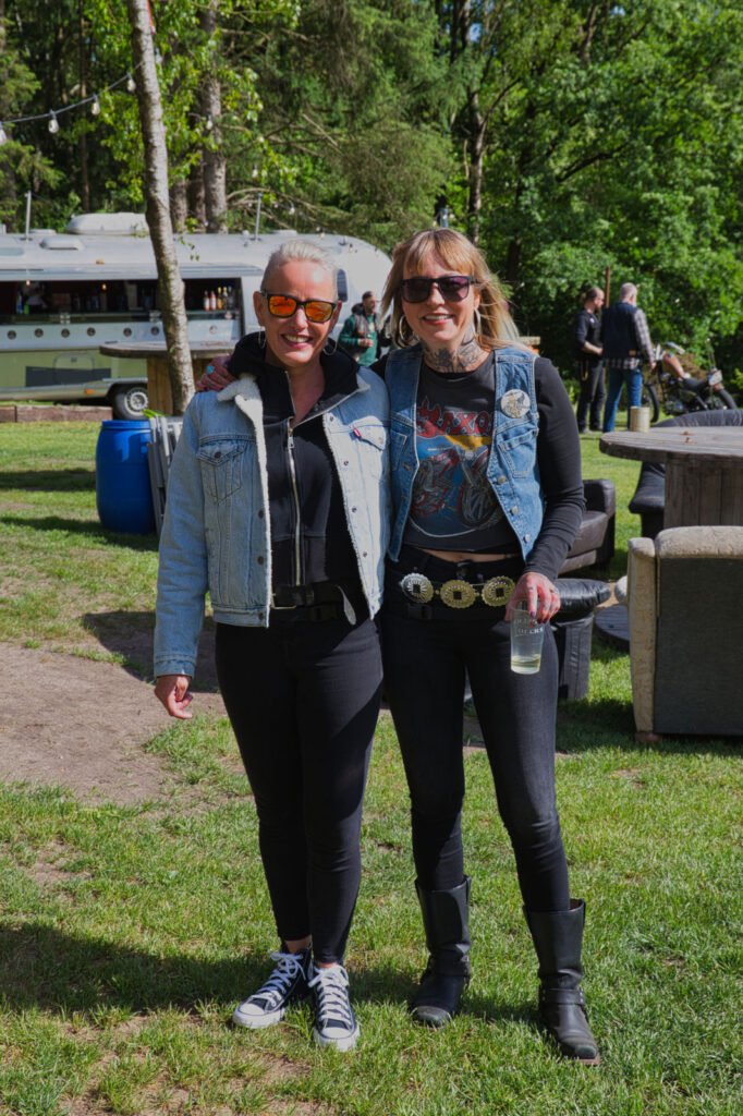 Zwei Frauen posieren und lächeln im Freien auf einer Wiese, beide tragen schwarze Hosen, helle Jeansjacken oder -westen, Sonnenbrillen und Freizeitschuhe. Im Hintergrund sind Bäume und Menschen zu sehen, und ein silberner Wohnwagen ist zu erkennen.