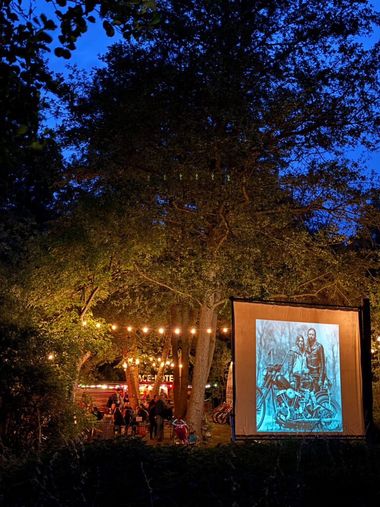Eine Filmnachtszene im Freien mit einem Projektorbildschirm, auf dem ein altes Foto von zwei Personen auf Motorrädern zu sehen ist; zwischen den Bäumen hängen Lichterketten, unter denen sich in der Abenddämmerung Menschen versammeln.