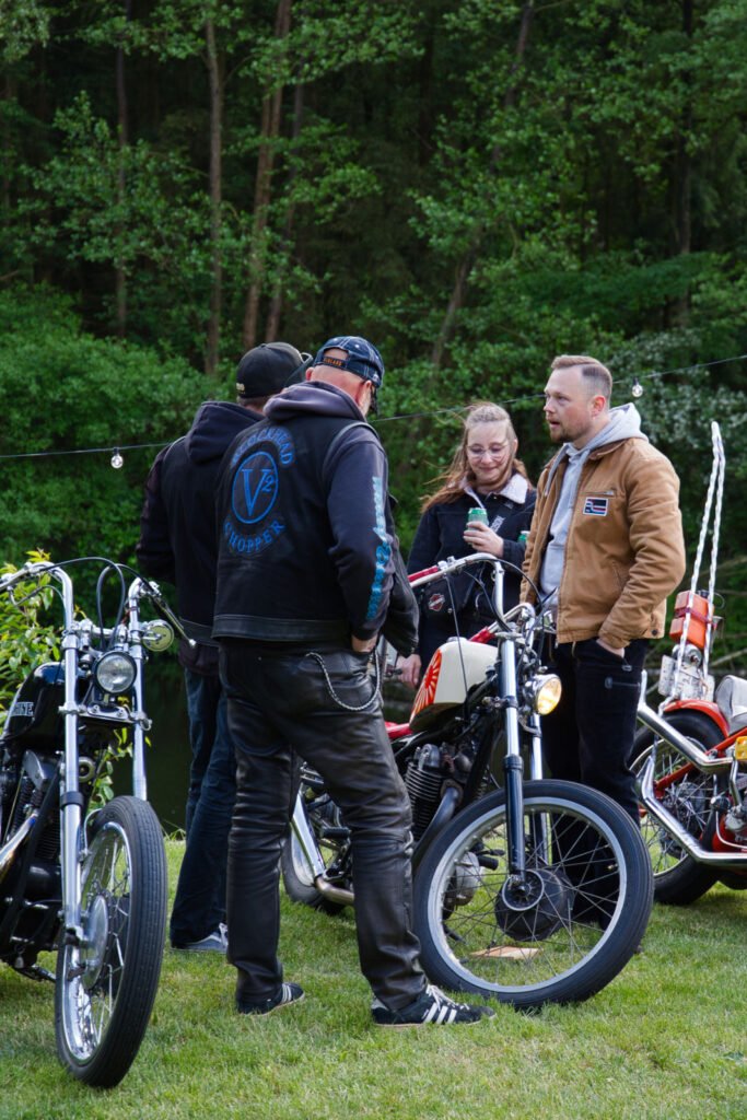 Vier Personen stehen im Freien zwischen alten Motorrädern, unterhalten sich und lächeln. Sie sind leger gekleidet, umgeben von grünen Bäumen und Gras, wobei zwei Motorräder im Vordergrund zu sehen sind.