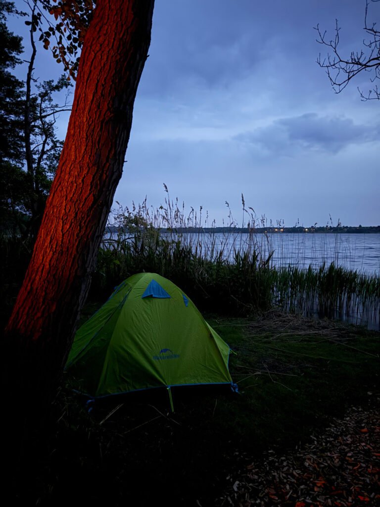 Ein grünes Zelt steht in der Abenddämmerung in der Nähe von hohem Gras an einem ruhigen See. Der Himmel ist bewölkt und ein Baum im Vordergrund wird von einem warmen, orangefarbenen Licht angestrahlt.