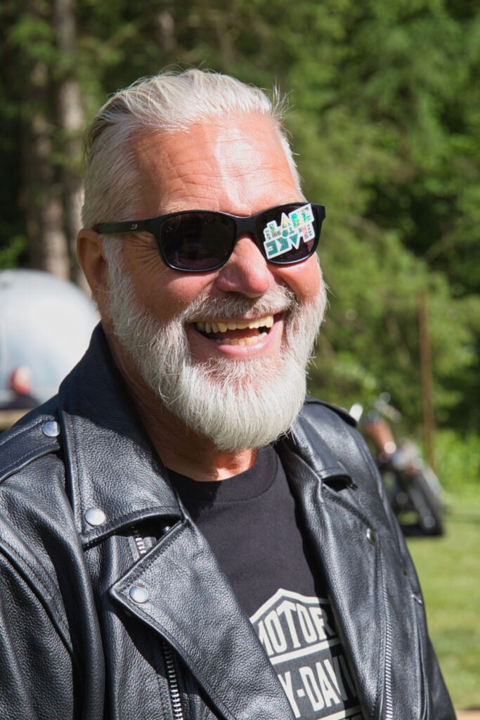 Ein lächelnder älterer Mann mit weißen Haaren und einem Bart trägt eine schwarze Sonnenbrille und eine schwarze Lederjacke im Freien. Auf der Spiegelung in seiner Brille steht "#LIVE". Im Hintergrund sind Bäume und Grünflächen zu sehen.