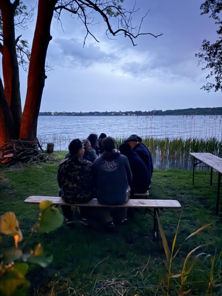 Eine Gruppe von fünf Personen sitzt eng beieinander auf einer Bank an einem See in der Abenddämmerung, umgeben von Bäumen und hohem Gras, mit Blick auf das Wasser und ferne Lichter am Ufer.