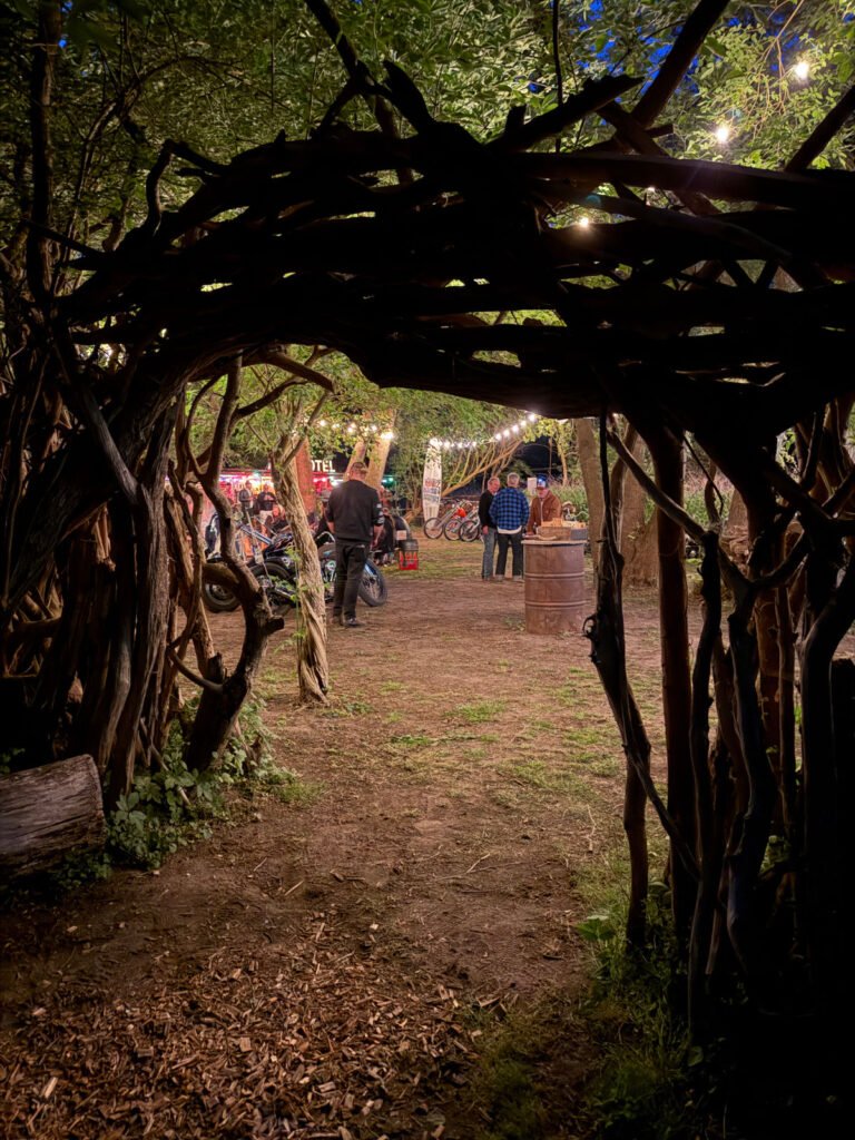 Ein hölzerner Torbogen aus gewundenen Ästen öffnet sich zu einer Lichtung, auf der Menschen stehen und sich unter Lichterketten unterhalten, umgeben von Bäumen und rustikaler Dekoration in der Abenddämmerung.