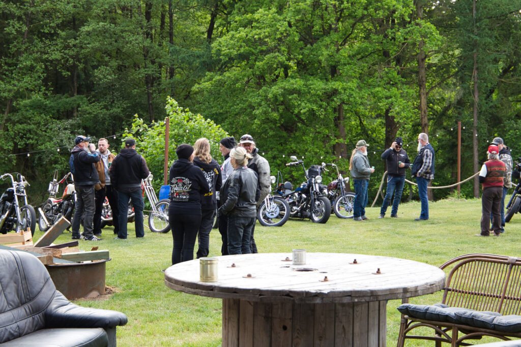 Eine Gruppe von Menschen, viele mit Lederjacken, versammelt sich im Freien in der Nähe mehrerer geparkter Motorräder auf einer von Bäumen umgebenen Wiese. Im Vordergrund stehen ein runder Holztisch und Gartenmöbel.