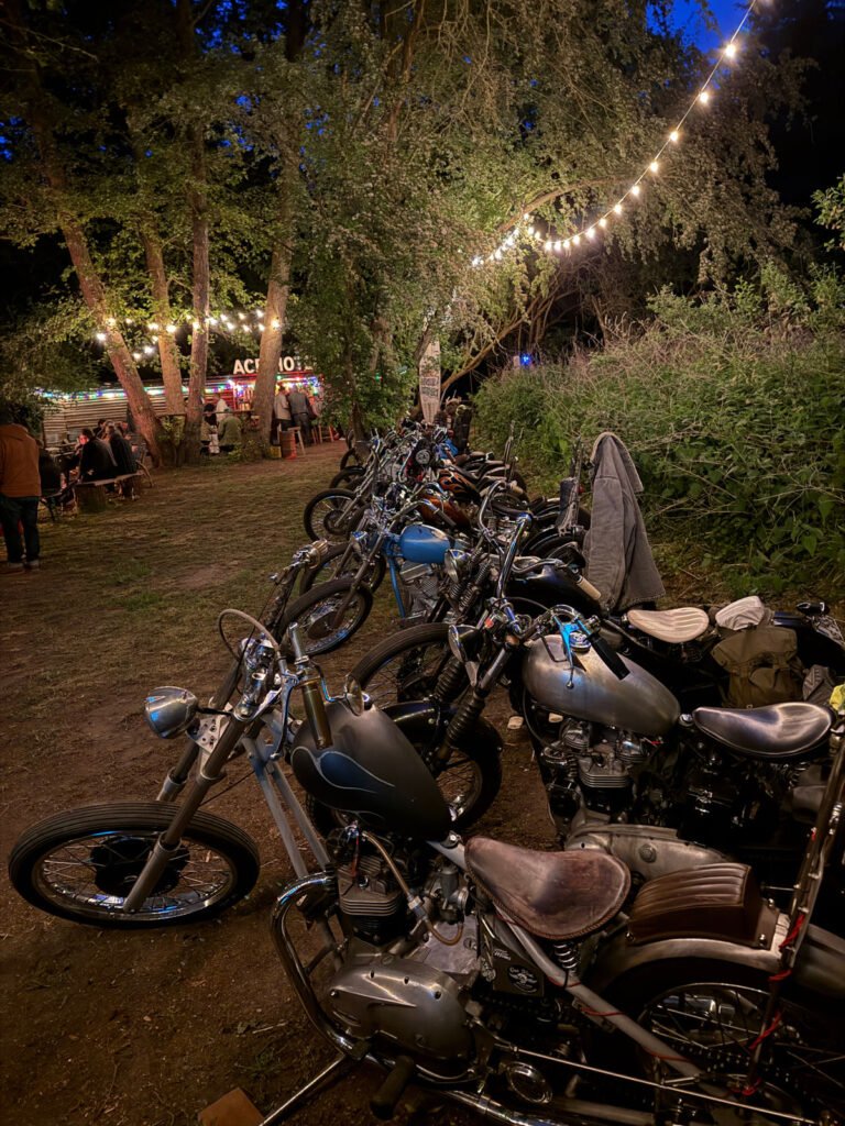 Eine Reihe von Oldtimer-Motorrädern ist nachts auf einer Wiese unter Lichterketten geparkt, im Hintergrund stehen Bäume und ein Essensstand, an dem sich Menschen versammeln.