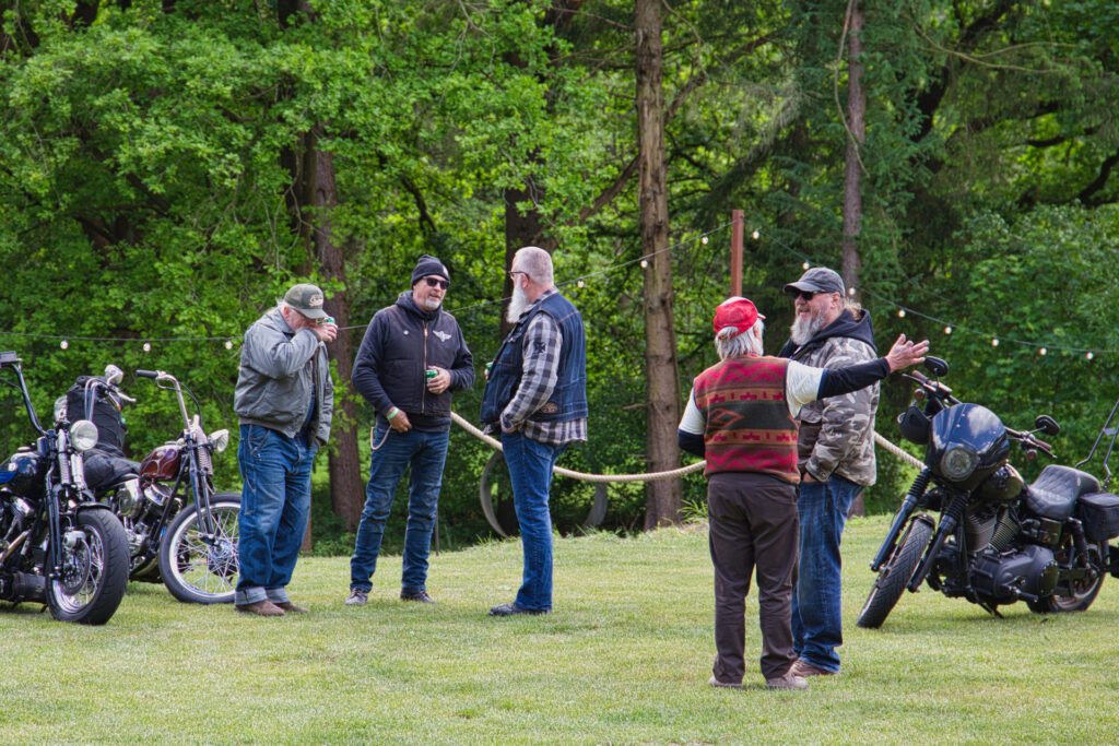 Fünf Männer stehen im Gespräch auf einer von Bäumen umgebenen Wiese, in deren Nähe mehrere Motorräder geparkt sind. Sie tragen legere Kleidung im Biker-Stil, wie Jacken, Westen und Hüte.