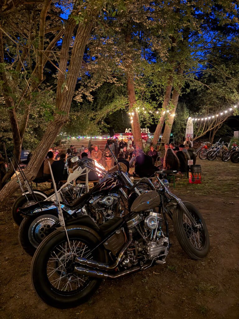 Mehrere Motorräder sind nachts unter Bäumen geparkt, während sich eine Gruppe von Menschen um eine Feuerstelle versammelt. Darüber hängen Lichterketten, die die Außenszene in der Nähe eines Gebäudes mit dem Neonschild "AC MOTEL" beleuchten.