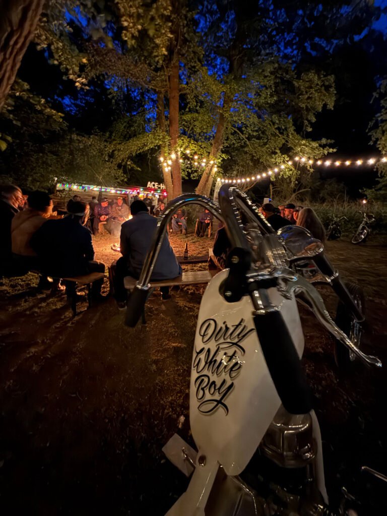 Eine Gruppe von Menschen sitzt nachts um ein Lagerfeuer unter Lichterketten, umgeben von Bäumen. Im Vordergrund ist ein weißes Motorrad mit der Aufschrift "Dirty White Boy" auf dem Tank geparkt.
