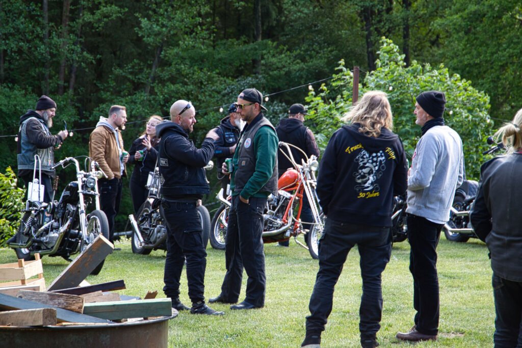 Eine Gruppe von Menschen in Motorradkleidung steht und unterhält sich neben geparkten Motorrädern auf einer Wiese, umgeben von Bäumen und Grün. Einige tragen schwarze Jacken mit Aufnähern und Logos.