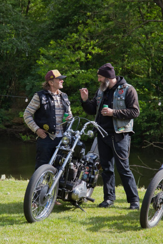 Zwei Männer in Motorradwesten und -hüten stehen neben einem Custom-Motorrad im Gras, halten Getränke in der Hand und unterhalten sich, während im Hintergrund Bäume und ein Fluss zu sehen sind.