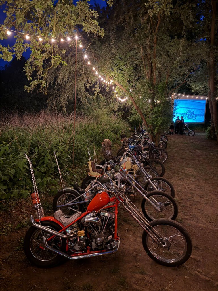 Eine Reihe von Custom-Motorrädern ist nachts auf einer Wiese unter hängenden Lichterketten geparkt, mit Bäumen und Büschen in der Nähe. Im Hintergrund versammeln sich Menschen vor einem großen Bildschirm, auf dem das Bild eines Motorrads zu sehen ist.