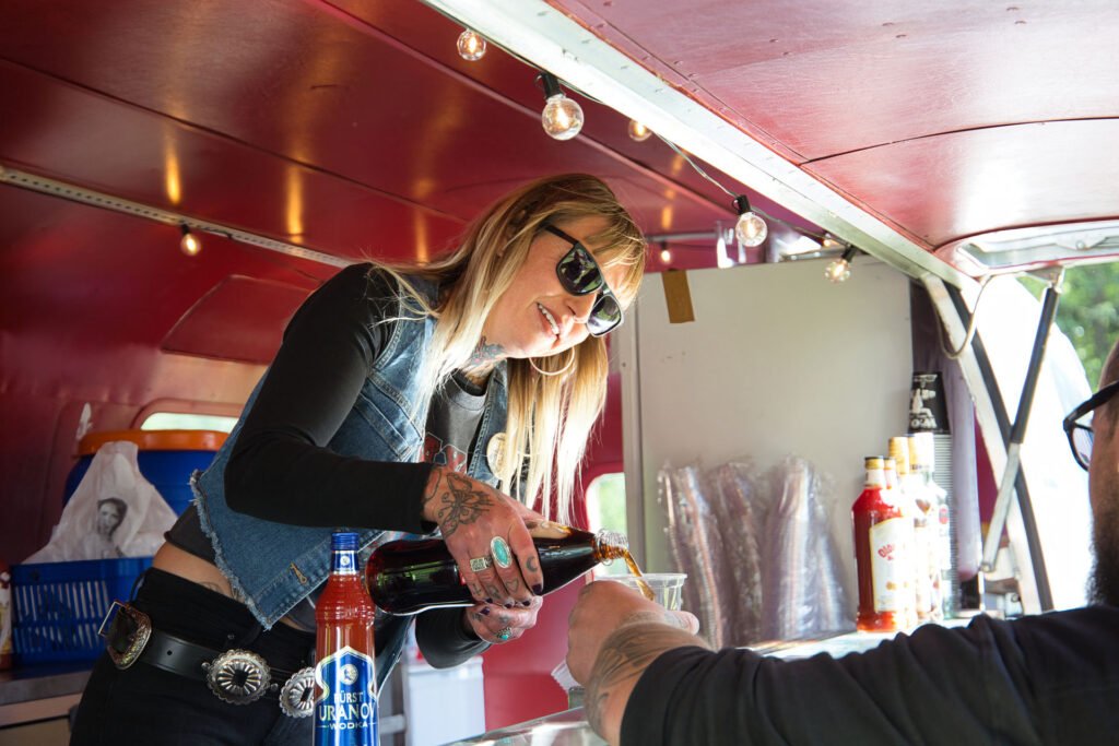 Eine Frau mit Sonnenbrille und Jeansweste schenkt an einem Imbisswagen ein Getränk in ein Glas ein und lächelt einen Kunden an. Auf dem Tresen neben ihr sind Flaschen und Becher zu sehen.