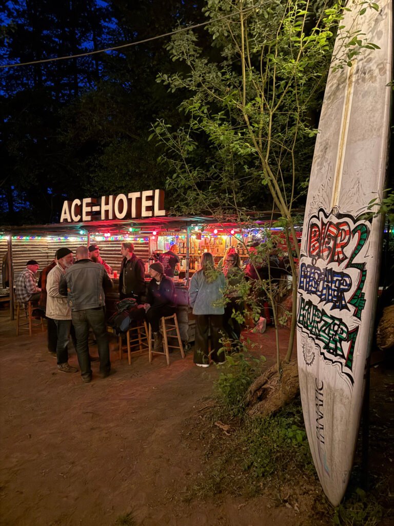 Eine Gruppe von Menschen trifft sich nachts an einer Bar im Freien unter Lichterketten. Die Bar ist mit einem "ACE HOTEL"-Schild versehen, und ein großes Surfbrett mit buntem Graffiti steht im Vordergrund zwischen Bäumen.