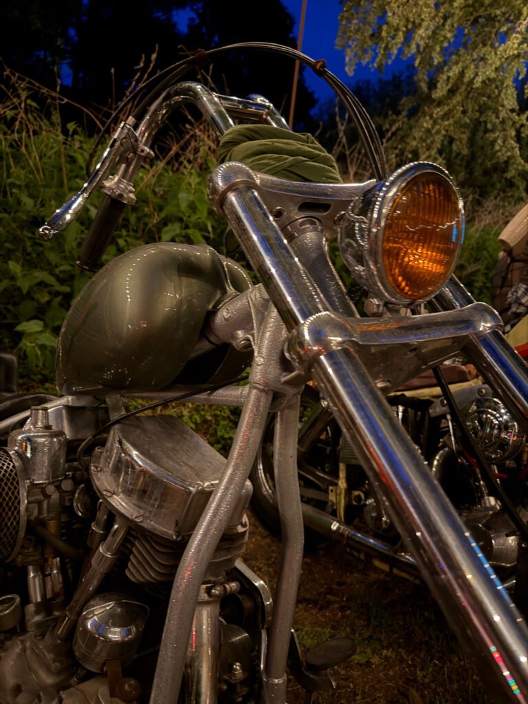 Nahaufnahme eines Oldtimer-Motorrads mit glänzendem Chromlenker, orangefarbenem Scheinwerfer und grünem Kraftstofftank. Das Motorrad ist im Freien in der Nähe von Grünflächen geparkt, mit Abendlicht im Hintergrund.
