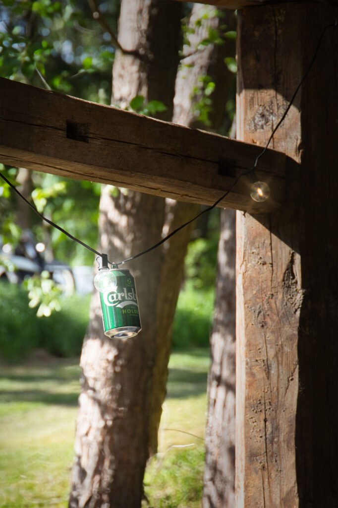 Eine grüne Carlsberg-Bierdose hängt an einer Lichterkette, die an einem Holzbalken befestigt ist, während im Hintergrund das Sonnenlicht durch Bäume und Grünzeug fällt.