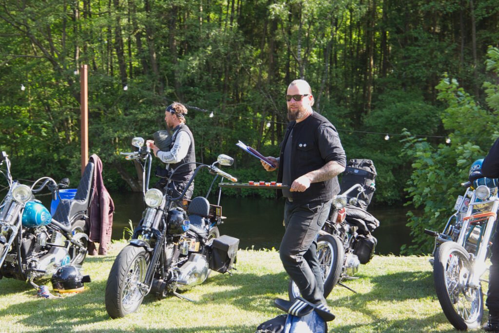 Zwei Männer stehen zwischen mehreren geparkten Motorrädern auf einer Wiese am Rande eines Waldgebiets. Ein Mann in Schwarz hält Papiere in der Hand und blickt in die Kamera, während der andere etwas an seinem Motorrad einstellt.
