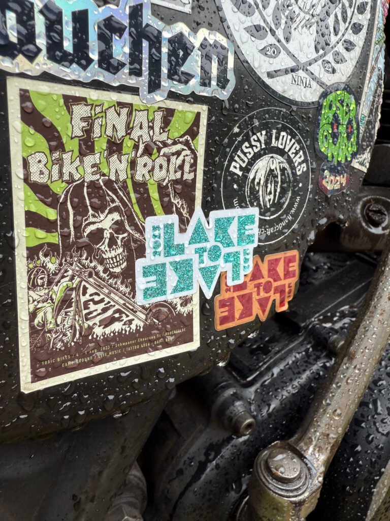 Nahaufnahme einer Metalloberfläche, die mit bunten Aufklebern bedeckt ist, darunter solche mit der Aufschrift "FINAL BIKE N' ROLL", "SLAVE TO SLAVE" und "PUSSY LOVERS!". Auf der Oberfläche sind Wassertropfen zu sehen.
