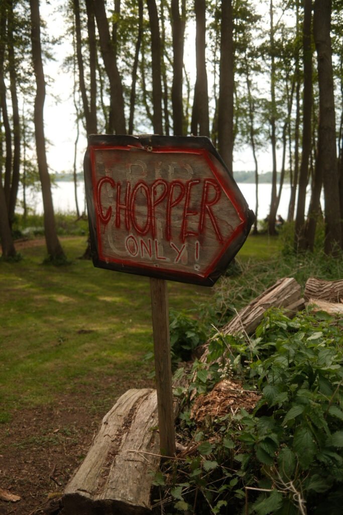 Ein handgefertigtes Holzschild mit roter Schrift "CHOPPER ONLY!" und einem nach rechts zeigenden Pfeil steht an einem Waldweg in der Nähe eines Sees, umgeben von Bäumen, Baumstämmen und Grünzeug.