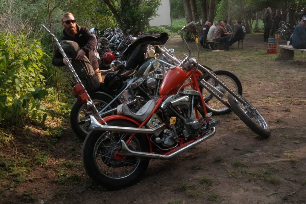 Ein bärtiger Mann mit Sonnenbrille steht neben einer Reihe von geparkten Custom-Motorrädern auf einem unbefestigten Weg, während im Hintergrund Menschen an Picknicktischen sitzen, die von Bäumen umgeben sind.