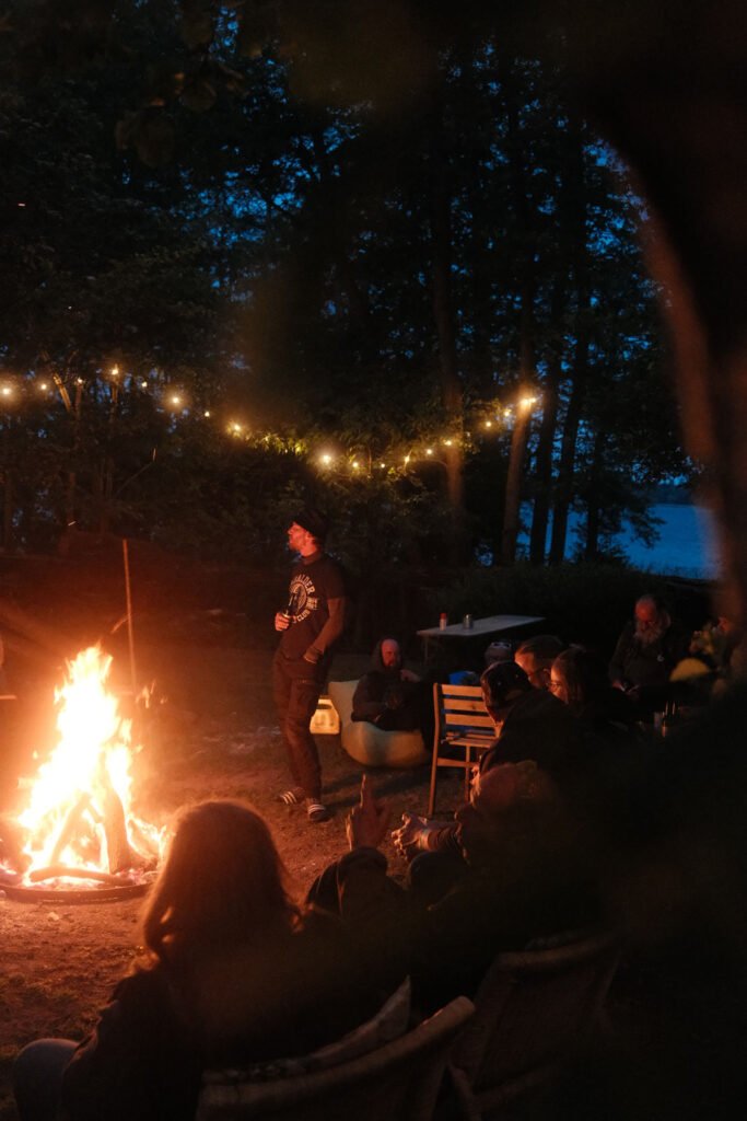 Eine Gruppe von Menschen sitzt in der Abenddämmerung um ein Lagerfeuer, umgeben von Bäumen und Lichterketten. Einige sitzen auf Stühlen, während eine Person am Feuer steht. Im Hintergrund ist ein See zu sehen.