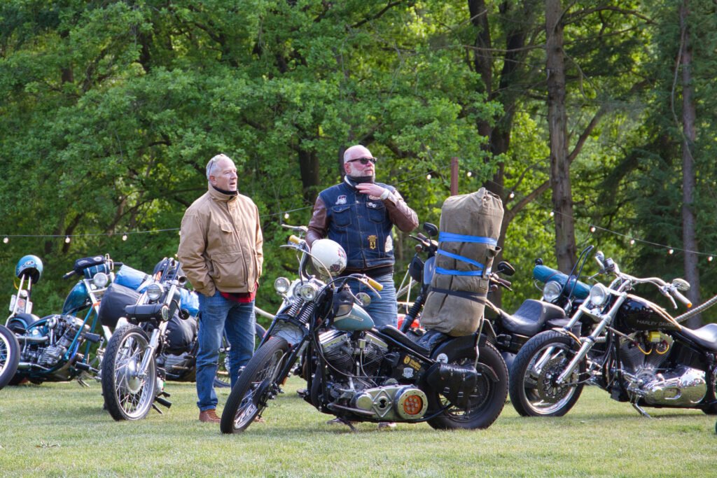 Zwei Männer stehen zwischen mehreren geparkten Motorrädern auf einer Wiese, mit Bäumen im Hintergrund. Ein Mann trägt eine hellbraune Jacke und Jeans, der andere eine schwarze Weste mit Aufnähern, eine Sonnenbrille und trägt einen großen Seesack auf seinem Motorrad.